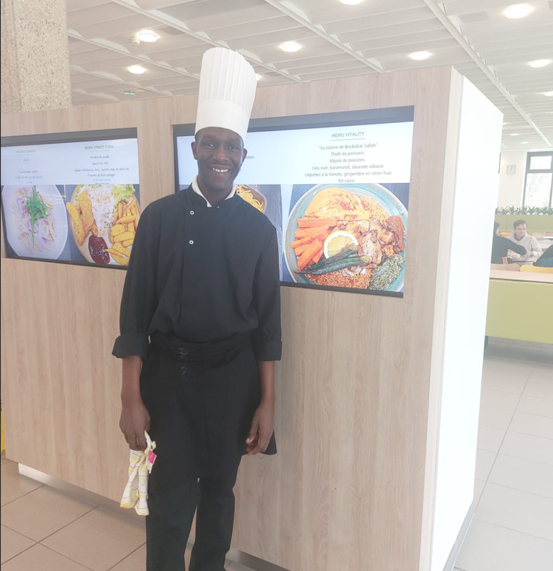 Boubakar Sallah, entrepreneur culinaire, en tenue de chef devant le menu « La cuisine de Boubakar Sallah » d'un restaurant Eldora